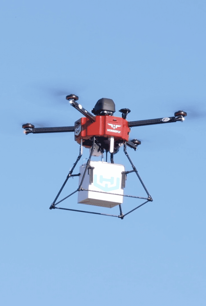  Photo Of Red Delivery Drone Carrying A White Package Against A Blue Sky.