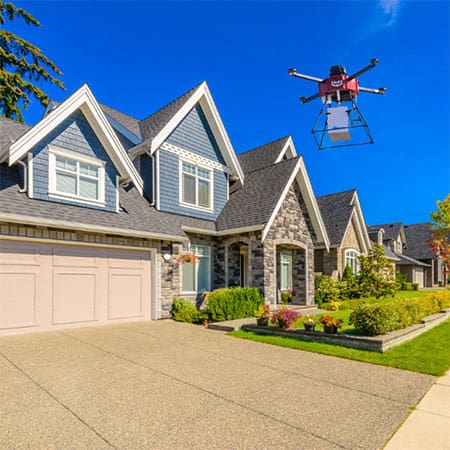 Drone with a package hovers over a suburban home's driveway on a sunny day