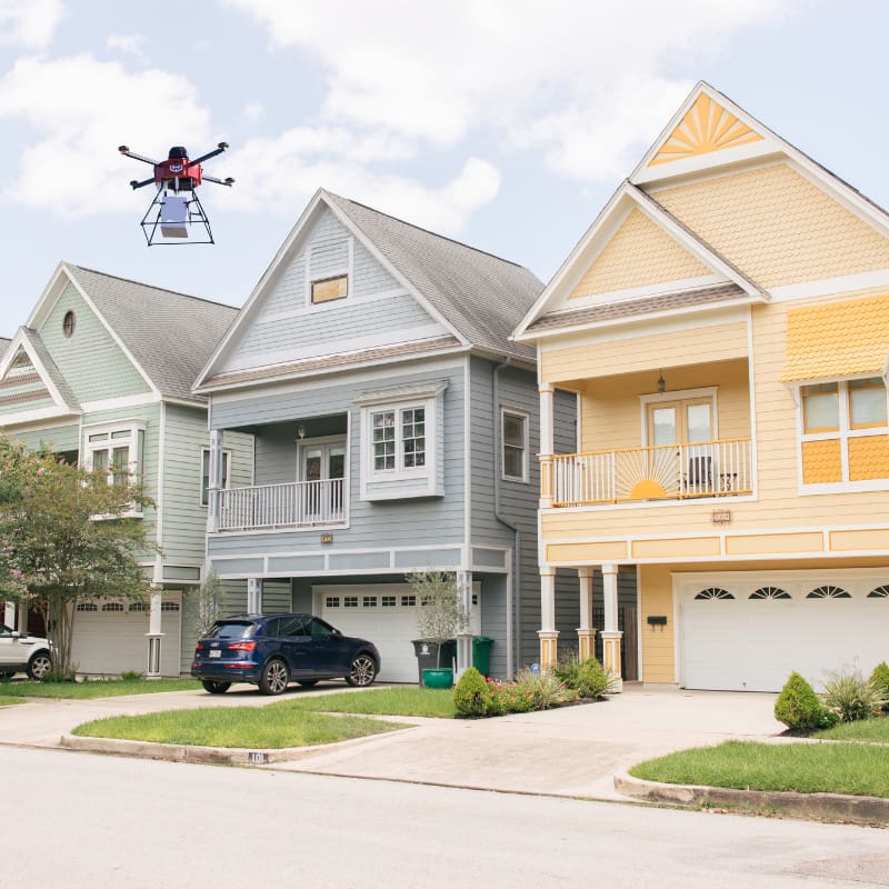 A Drone With A Package Hovers Above A Suburban Street With Colorful Two-Story Houses And Parked Cars On A Sunny Day.