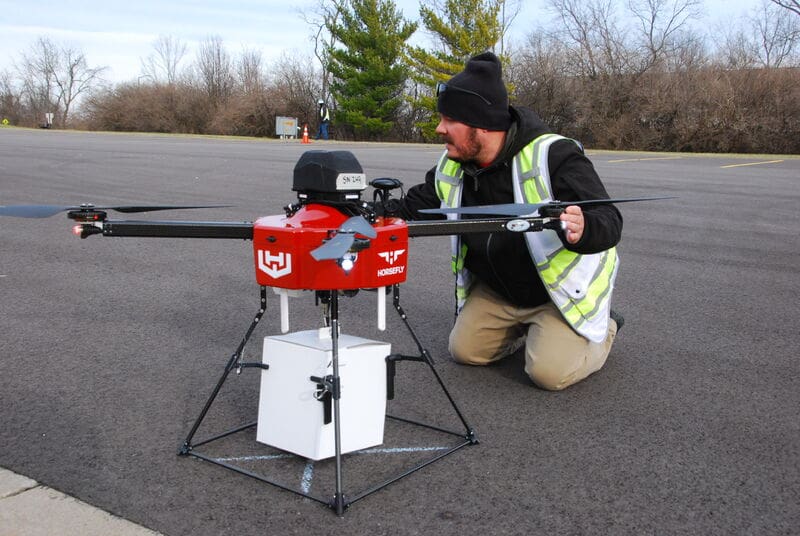 Technician Kneels To Adjust A Red Drone On The Ground With A Cargo Box Attached.
